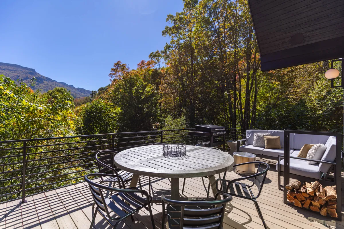 An outdoor dining space featuring a circular table and comfortable seating is set on a deck overlooking the surrounding trees and distant mountain views. The setting suggests social gatherings, relaxation, and immersive nature experiences.