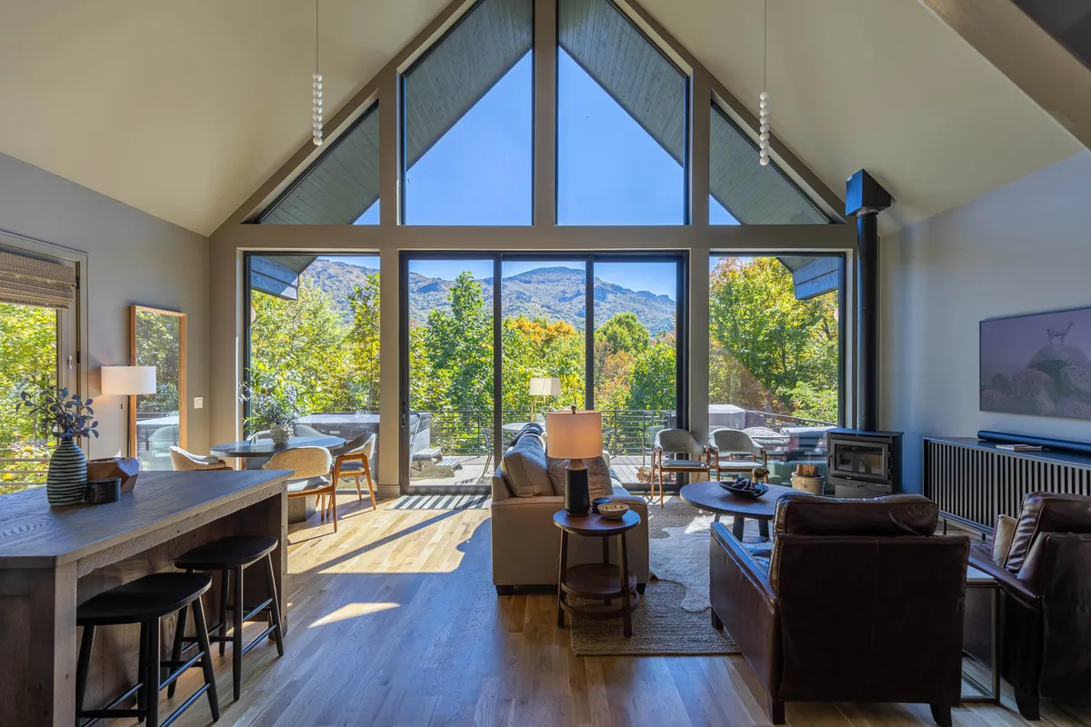 This image features a spacious living area with large windows offering breathtaking mountain views, a comfortable seating area, and a wooden bar counter. The abundant natural light and the direct sightlines to the forested landscape evoke the feeling of being surrounded by nature even while indoors.