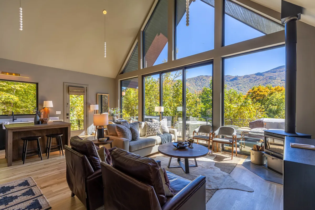 A spacious great room with comfortable seating oriented toward a modern fireplace, large windows framing sweeping mountain views, and an adjacent dining area. The room is filled with natural light and offers an inviting atmosphere for gatherings or quiet relaxation.