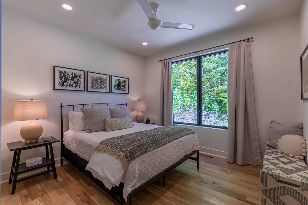 A serene bedroom with a plush bed, large window letting in natural light, a nightstand with a lamp, and tasteful wall art. The linens appear soft and inviting, and the overall ambiance is peaceful and restful.