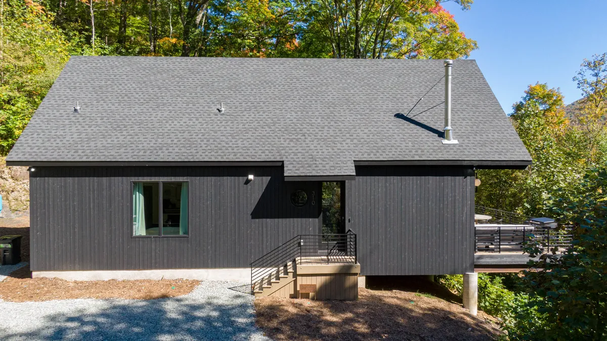 A contemporary cabin with sleek black exterior nestled among lush greenery. The spacious deck area is set up for outdoor gatherings, with surrounding trees hinting at nearby hiking trails and natural exploration.