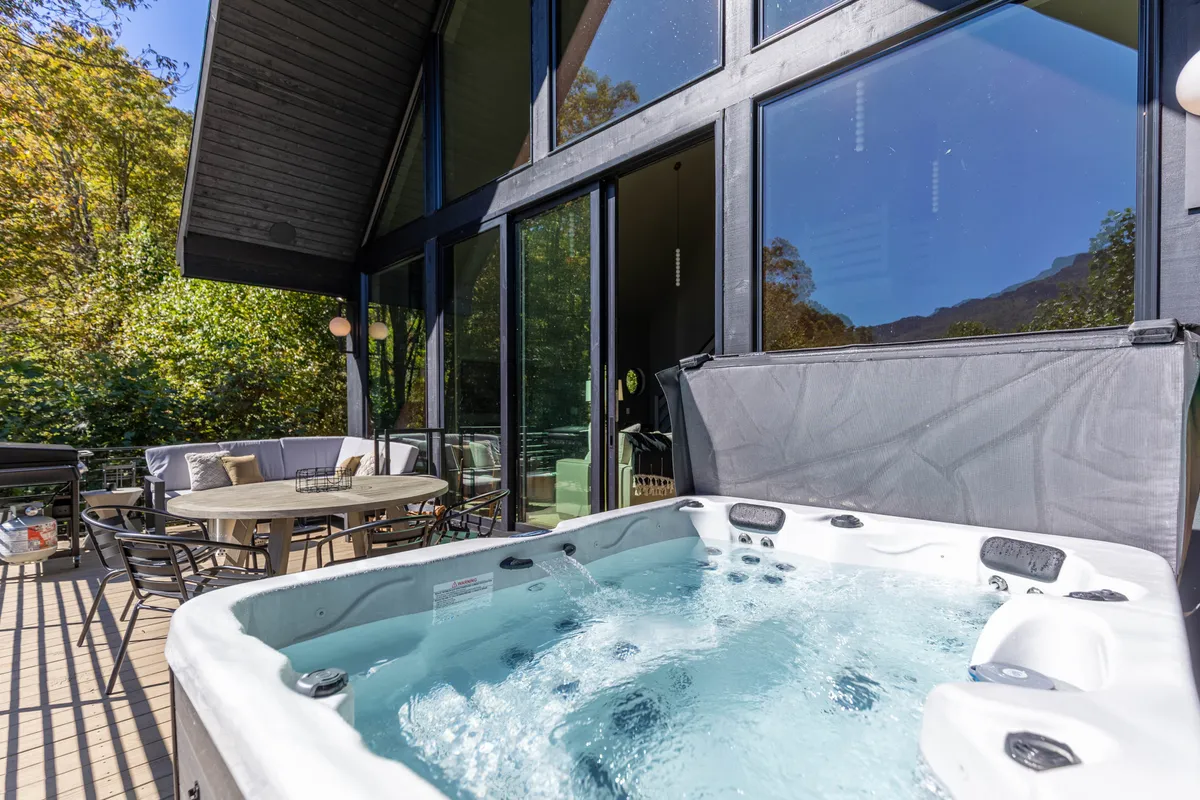 A tranquil hot tub set on a wooden deck, surrounded by an outdoor seating area and large glass windows that reflect the surrounding scenery. The atmosphere is peaceful, with hints of sunset light, inviting guests to soak and relax while taking in the views.