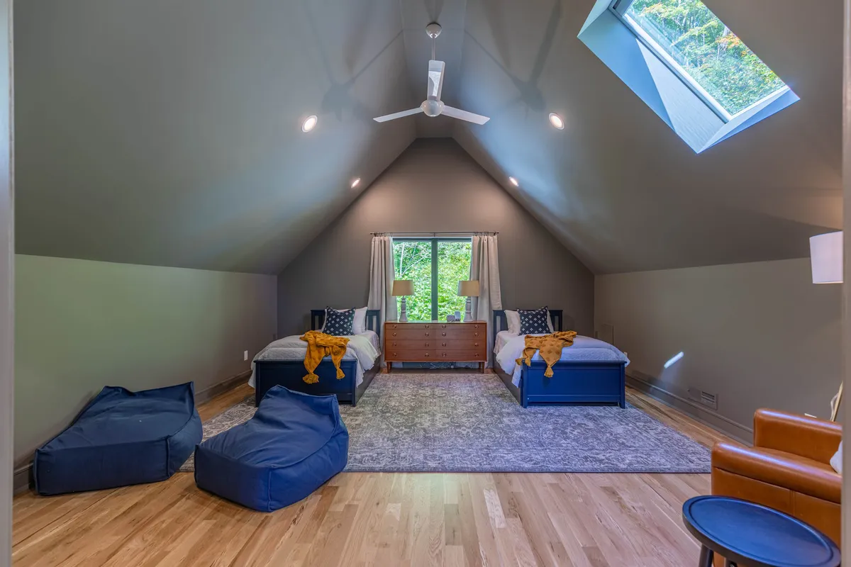 A playful loft bedroom featuring two twin beds, a large window, a skylight, bean bag chairs, and a wooden dresser. The space feels bright and welcoming, ideal for kids or friends to unwind, play, and relax.