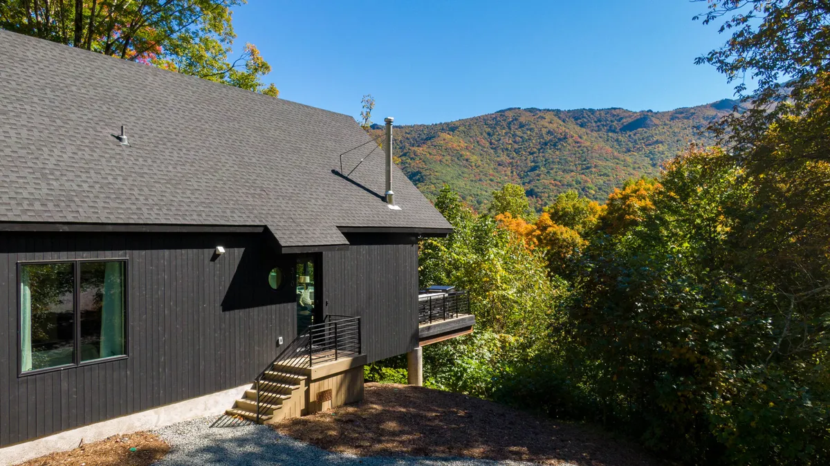 A striking exterior shot of the cabin, featuring its modern architecture nestled among tall trees with a mountain backdrop. The spacious deck is visible, ideal for outdoor gatherings and relaxation, while the surrounding landscape offers ample room for pets to explore and play.