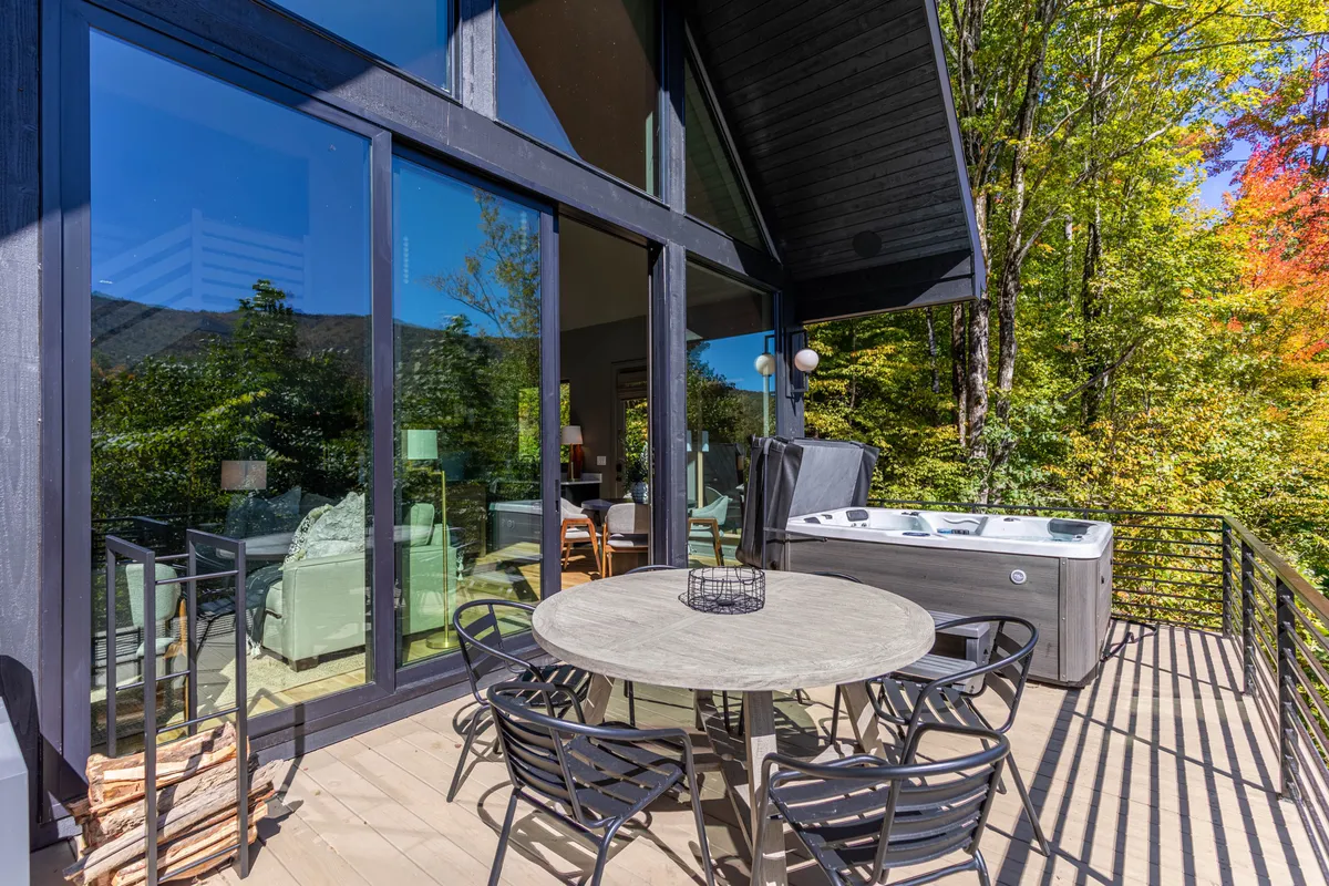This image presents a spacious deck featuring an outdoor dining table and hot tub, with large glass windows in the background and lush greenery all around. The scene captures both the relaxation opportunities in the hot tub and the potential for al fresco dining, with the deck seamlessly blending into its natural surroundings.