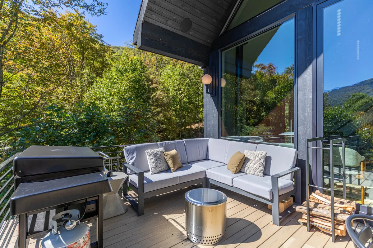 A comfortable outdoor seating area featuring a sectional sofa, a grill ready for barbecuing, and a fire pit, all set against a backdrop of greenery. The scene is set for social gatherings and relaxation, with the potential for enjoying sunset views and cozy fireside chats.