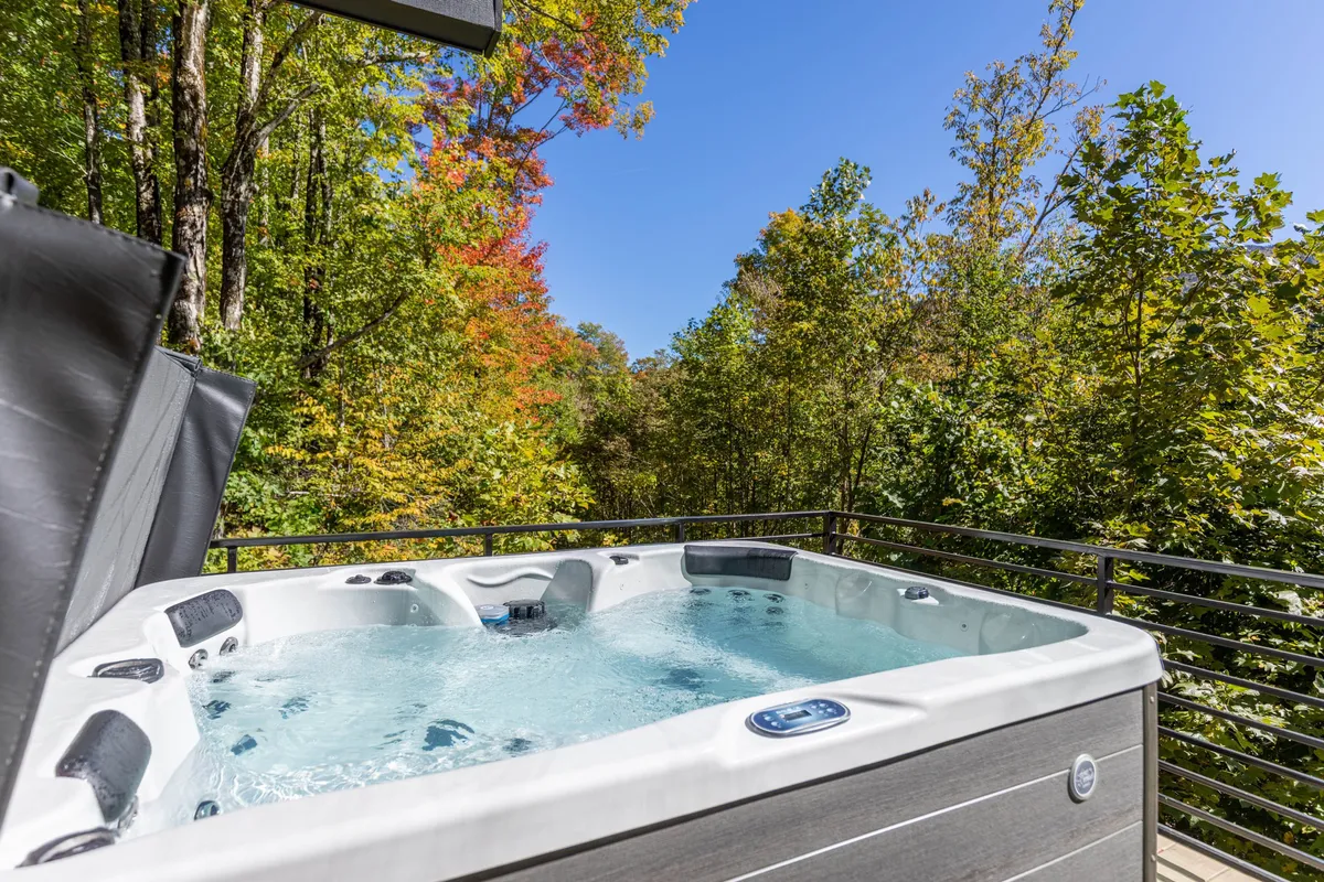 A tranquil hot tub sits amidst tall trees under a clear sky. The natural surroundings give a sense of seclusion and peace, with hints of foliage and the promise of stargazing or relaxation after outdoor adventures.