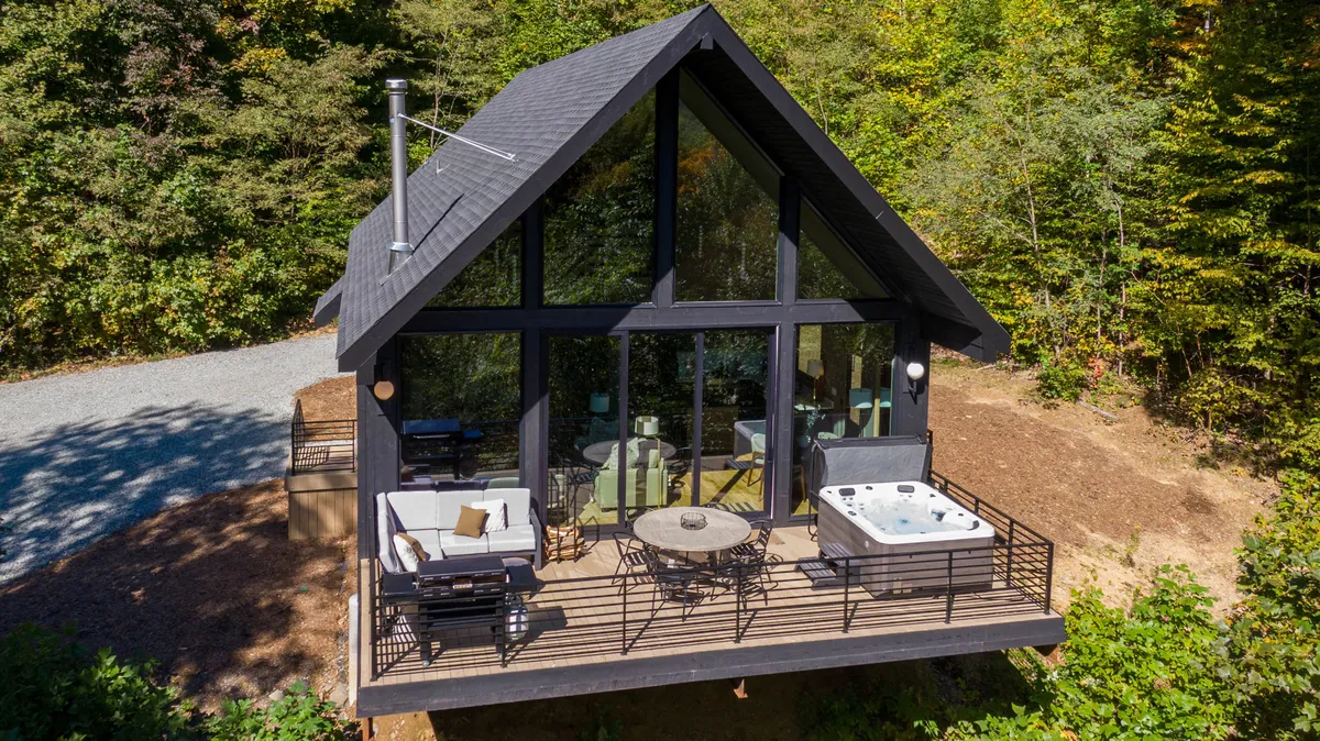 The image showcases the striking exterior of a modern luxury cabin nestled among the trees, featuring expansive windows that invite natural light, a sparkling hot tub on the deck, and inviting outdoor seating. The tranquil forest surrounds and mountain backdrop create a sense of peaceful seclusion, while the deck is set up for both relaxation and social gatherings.