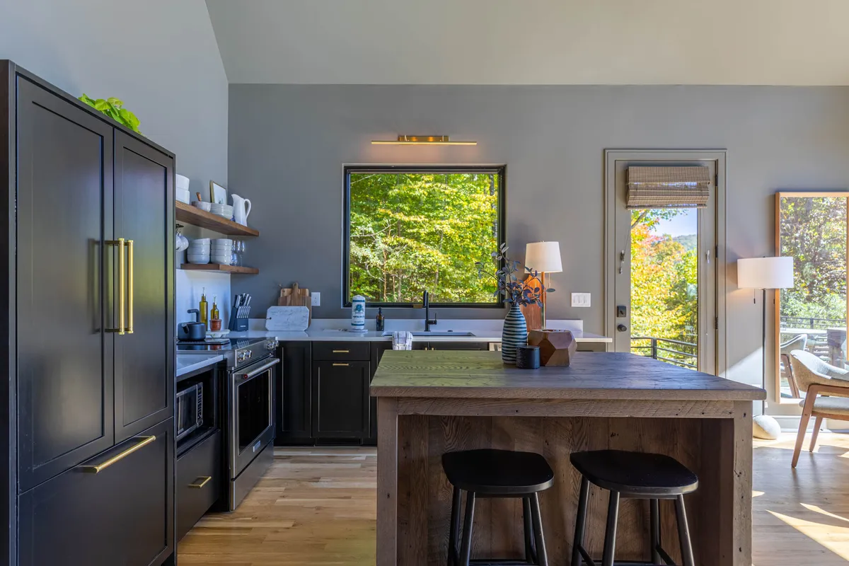 A spacious, open-concept kitchen featuring a large wooden island, sleek black cabinetry, and a generous window offering a view of trees outside. The setup is perfect for group cooking, with ample space for entertaining and gathering.