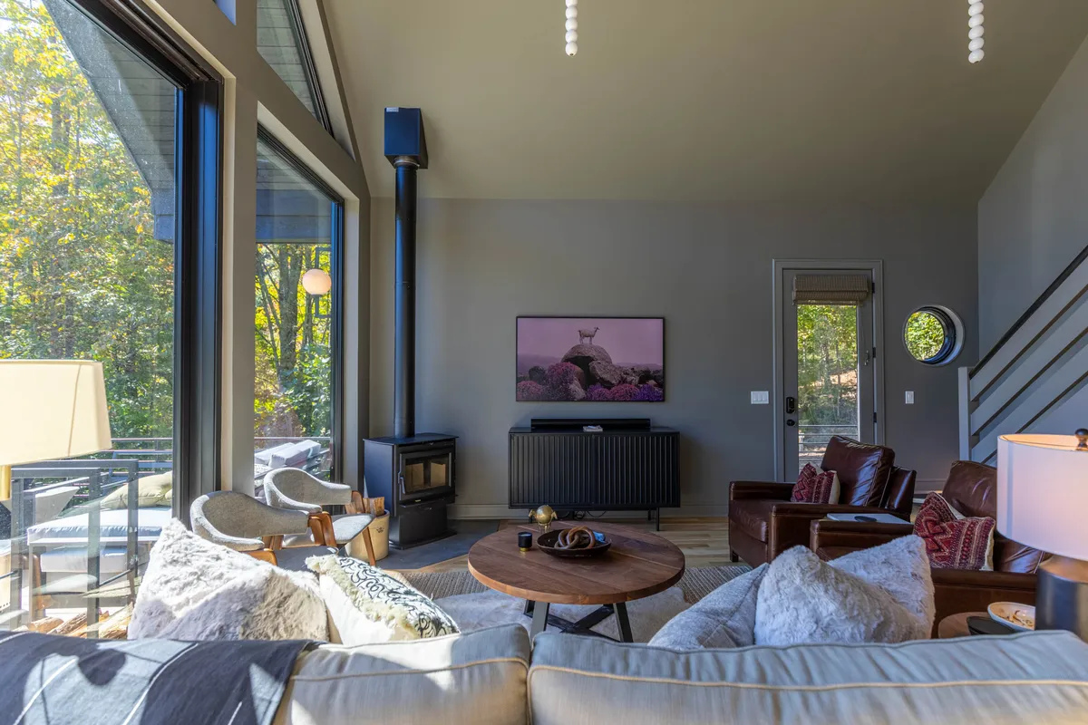 A cozy living area featuring a wood-burning stove, modern television, ample seating arrangements, and floor-to-ceiling windows that showcase beautiful sunset-lit mountain vistas. The round coffee table and decor foster an inviting and sociable setting.